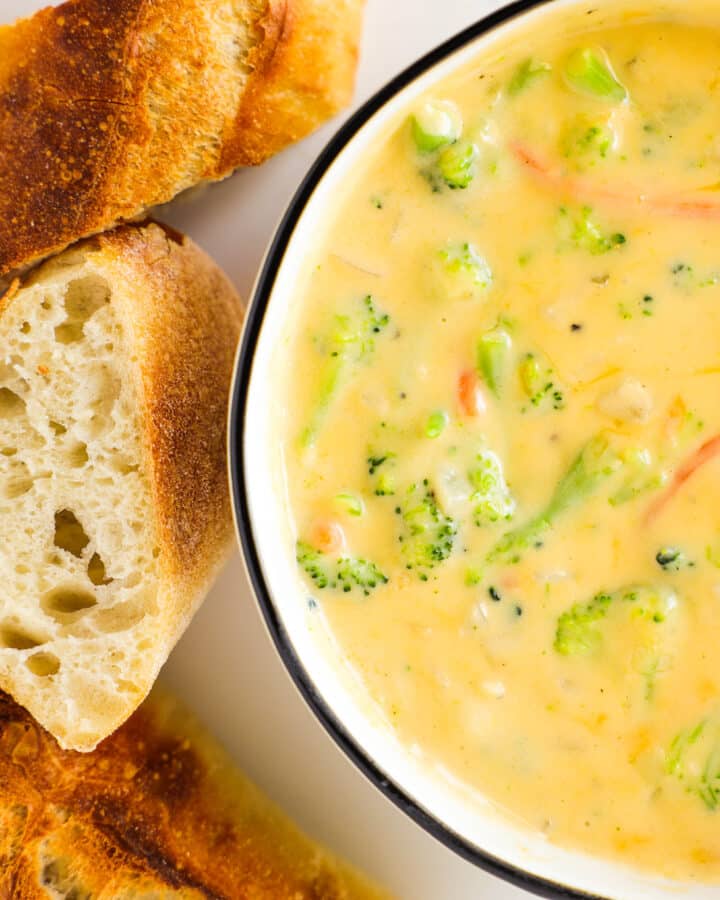An overhead view of a bowl of Broccoli Cheese Soup with slices of baguette beside it.
