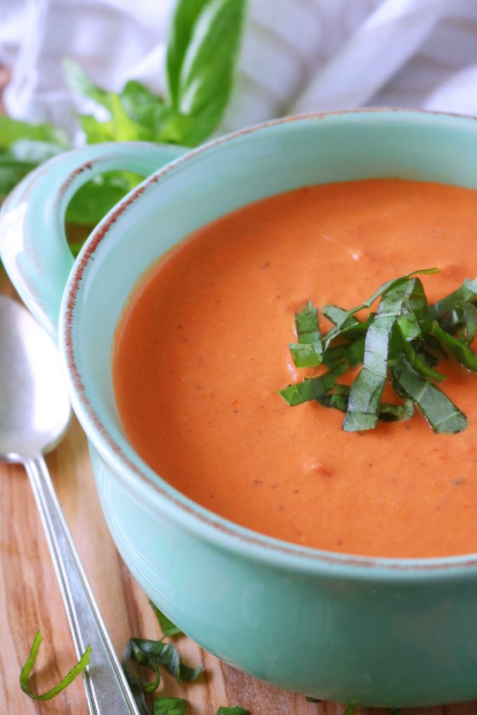 A close up shot of homemade tomato soup served in a light blue bowl with slivers of fresh basil in the center of the bowl. Lying flat on the left side of the bowl is a silver spoon. 