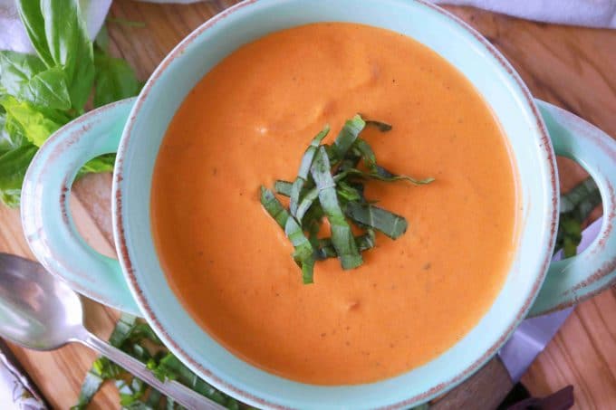 An overhead shot of homemade tomato soup served in a light blue bowl with handles. In the center of the bowl of soup are slivers of fresh basil. 