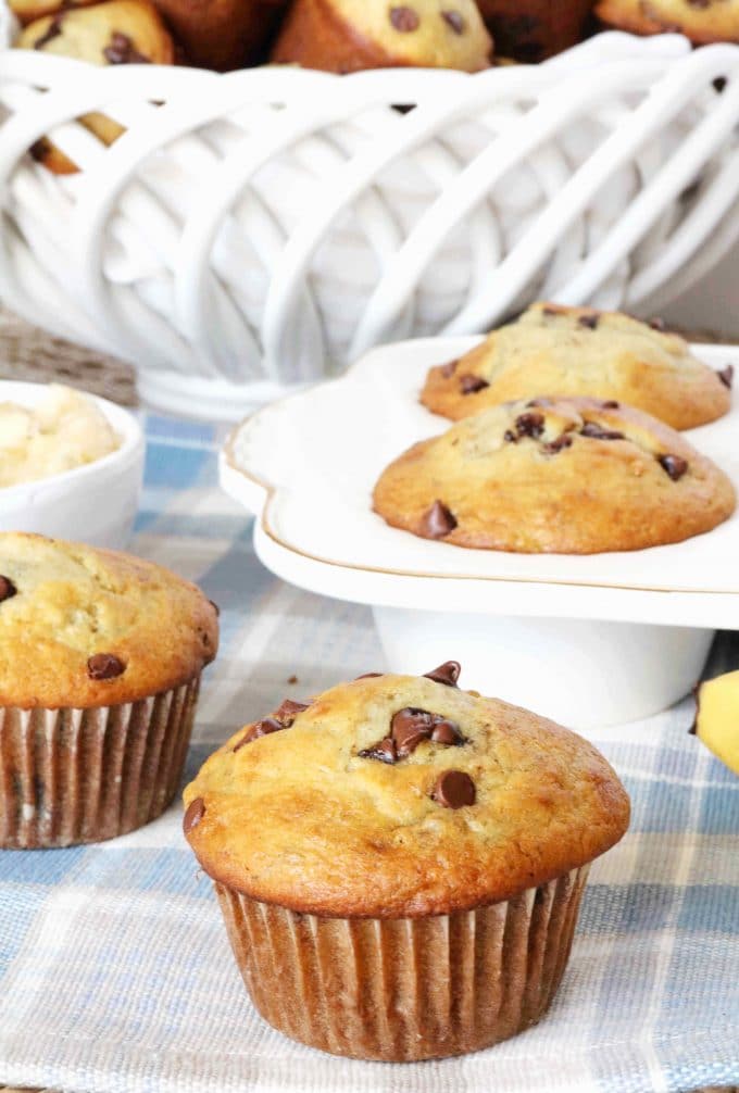 A close up shot of banana and chocolate chip muffins sitting on top of a white and blue plaid tablecloth. Behind the muffins are a white muffin tray with muffins in the them. 