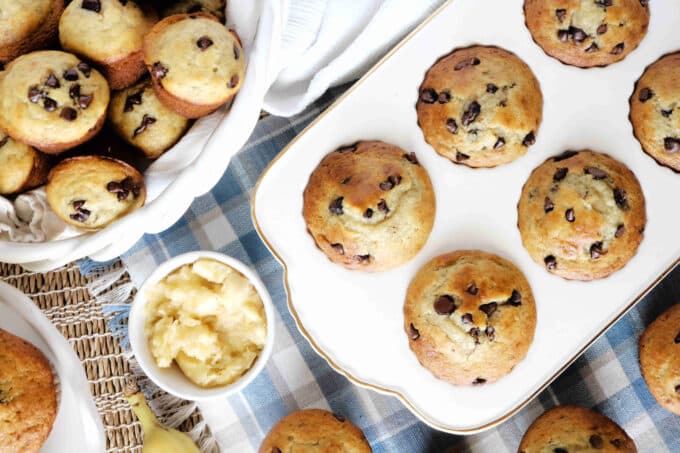 An overhead shot of banana and chocolate chip muffins in a white stone muffin tray. Next to the muffin tray is a bread basket full of muffins. There is also a small white bowl of mashed banana. 