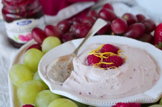 Fruit dip in a white serving bowl with a spoon resting on the side of the bowl. In the center of the dip are three raspberries topped with lemon zest. Surrounding the bowl are red and green grapes. In the background is a jar of raspberry preserves. 