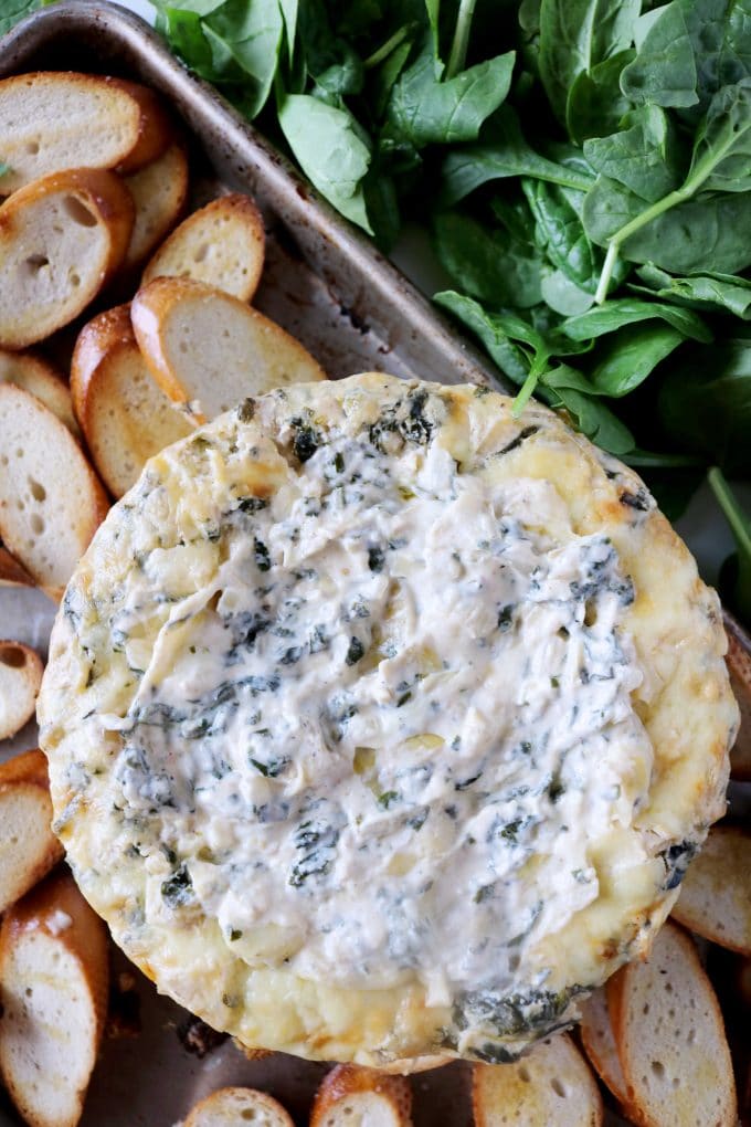 An overhead shot of spinach artichoke dip surrounded by toasted baguettes and fresh spinach leaves. 