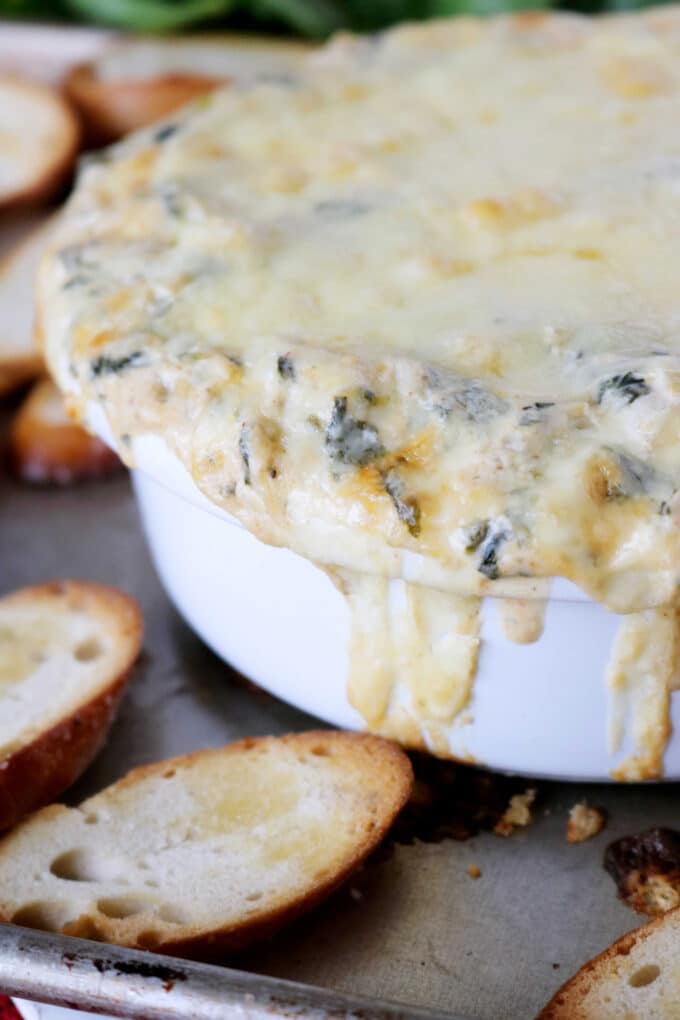 A side view of Spinach and Artichoke Dip that is sitting on top of a baking tray surrounded by toasted baguettes. 