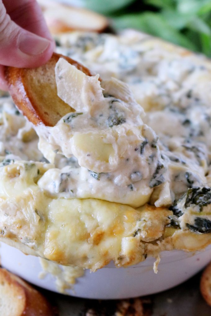 A hand dipping a baguette in to the Spinach and Artichoke Dip. 