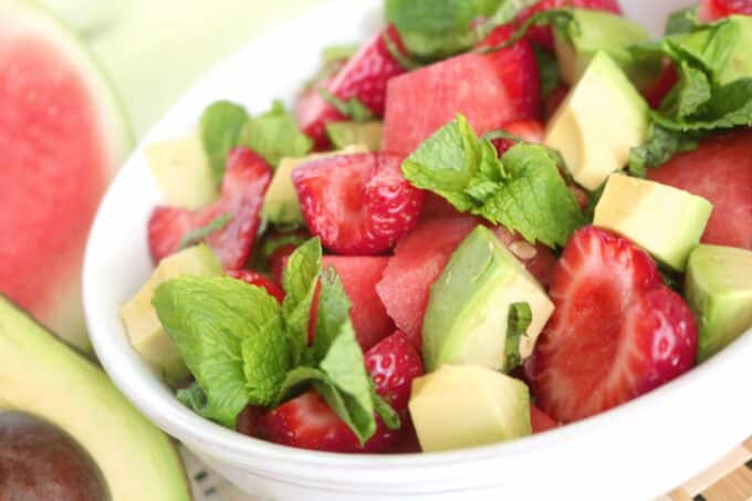 Watermelon and strawberry salad in a white serving bowl. Off to the right is a halved avocado and watermelon. 