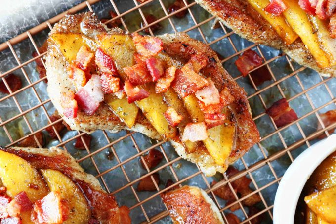 An overhead shot of pork chops topped with apples and bacon resting on a wire cooling rack. 