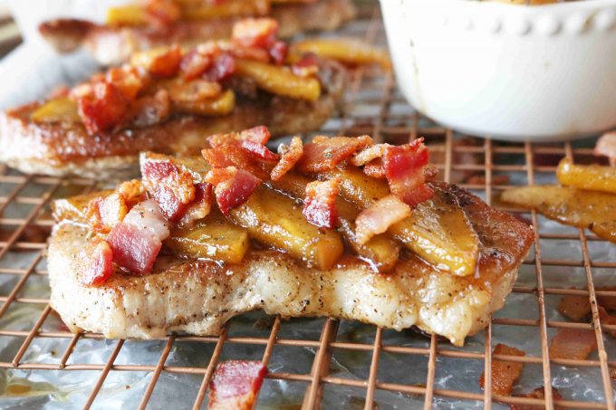 Cooked pork chops topped with apples and bacon resting on a wire cooling rack. 