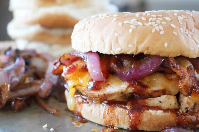  A close up shot of BBQ chicken breast sandwich layered with purple onions, cheese, chicken, bbq sauce and a sesame bun. 
