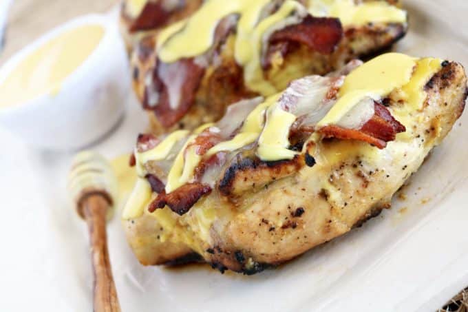 Two chicken breast layered with bacon, cheese and honey mustard sauce sitting on top of a white serving dish. Off to the side is a wooden honey spoon and a bowl of the honey mustard sauce. 