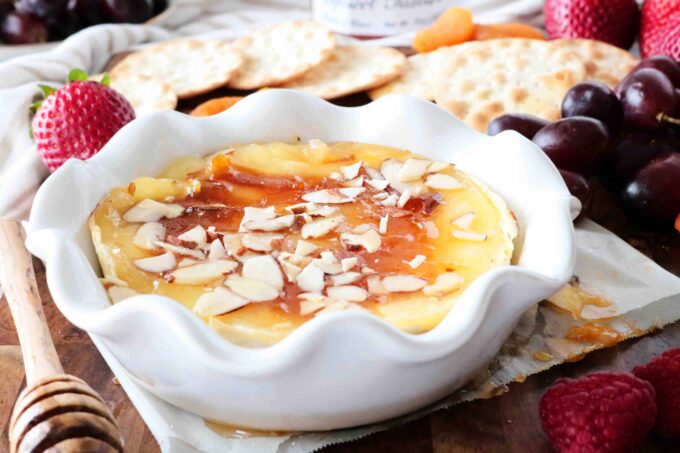 Baked brie topped with jam and chopped almonds on a wooden board surrounded by strawberries, grapes, and water crackers.