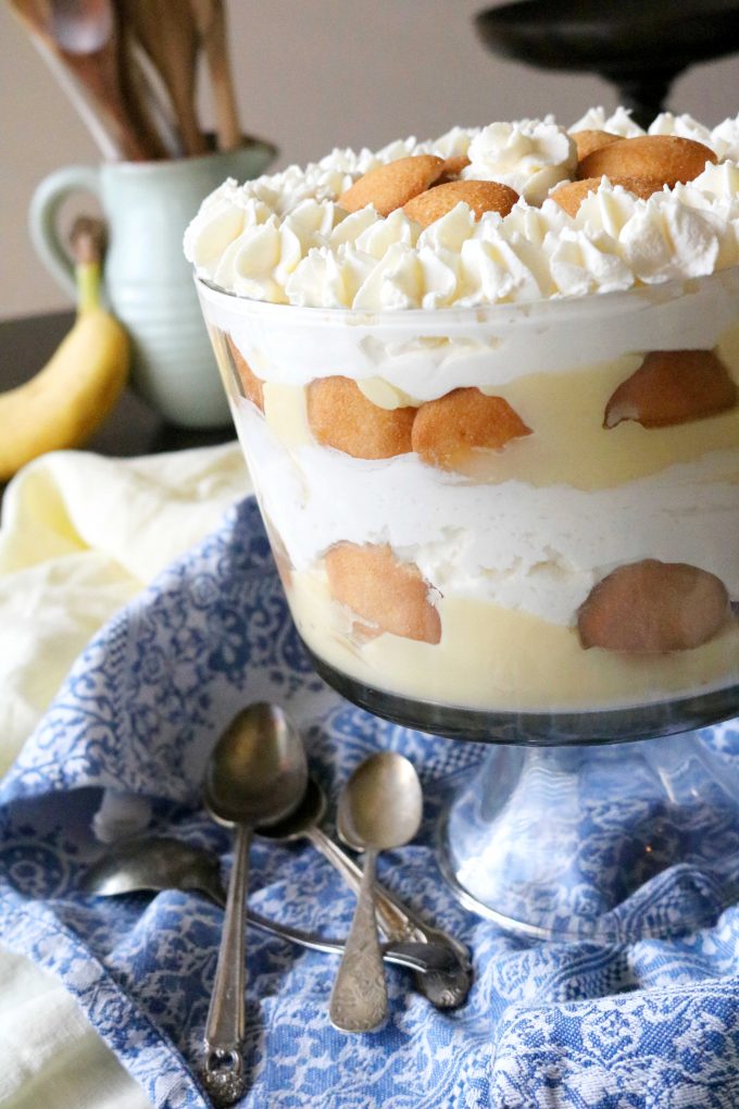Banana Pudding Trifle in a glass trifle bowl layered with banana pudding, Nilla Wafers and whipped cream. The bowl is sitting on top a blue and white printed napkin. 