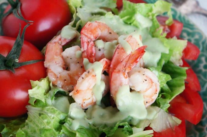 An overhead shot of shrimp sitting on top of romaine lettuce that has been drizzled with green goddess dressing. At the top left corner are whole fresh tomatoes. 