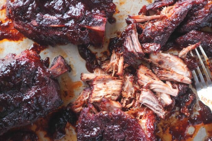 A close up shot of four country-style pork ribs. One of the ribs is shredded with a fork lying next to it. 