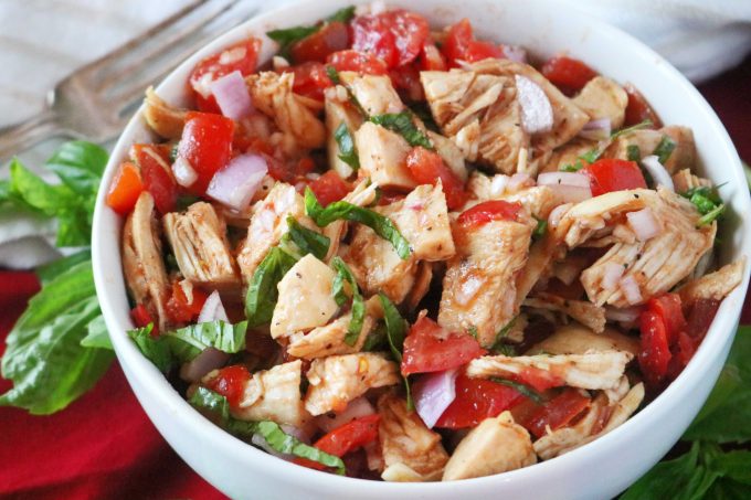 Sliced basil, tomatoes, purple onion, and cooked chicken all mixed together in a white bowl. In the background are whole basil leaves and a silver fork. 