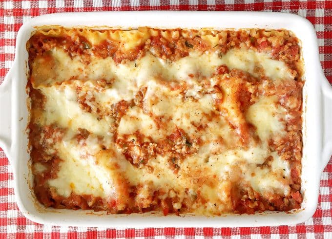 The lasagna bolognese is in a white baking dish that is sitting on top of a red and white checkered tablecloth. 