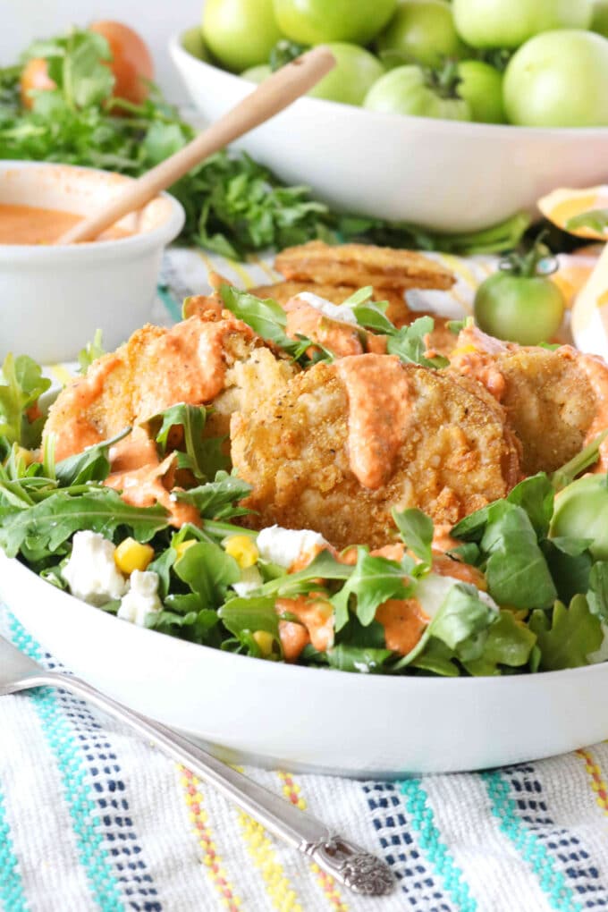 Fried green tomato salad being served in a white bowl. Behind the bowl of salad is a small white bowl of salad dressing with a wood spoon resting in it. 