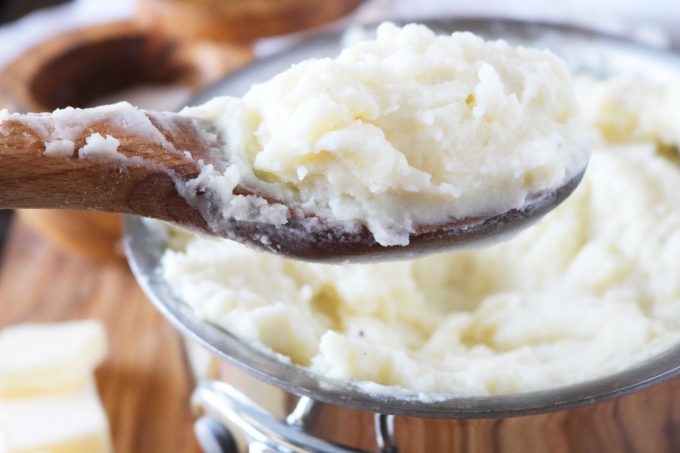 A wood spoon full of mashed potatoes being held over a pot full of mashed potatoes. 
