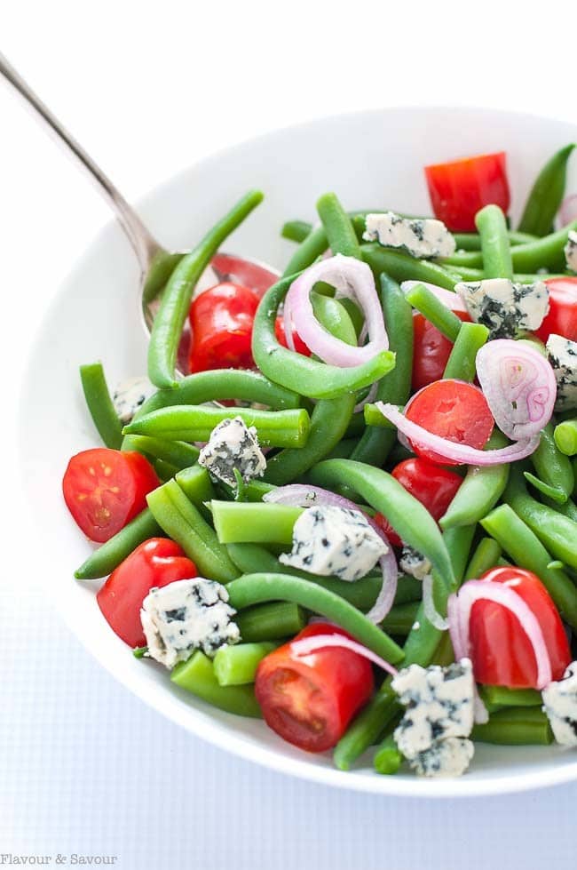 A close up shot of green beans, cherry tomato halves, sliced red onions and chunks of blue cheese. 