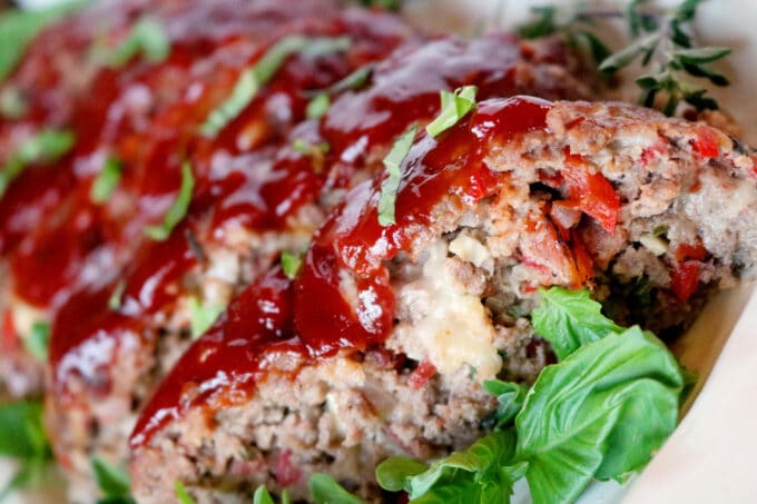 A close up shot of Italian Meatloaf on a white serving dish. Fresh basil is sprinkled on top and lying next to the meatloaf. 