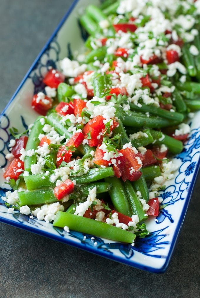 Green beans with tomatoes and feta cheese being served on a blue and white rectangle plate. 