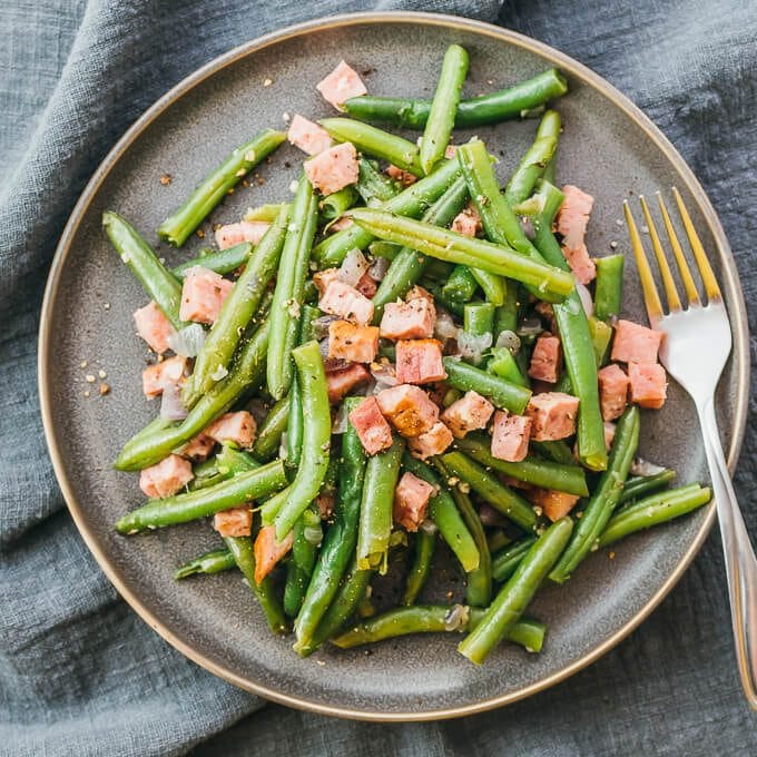 An overhead shot of green beans topped with cubed ham all served on a dark gray plate with a fork lying next to the beans. 