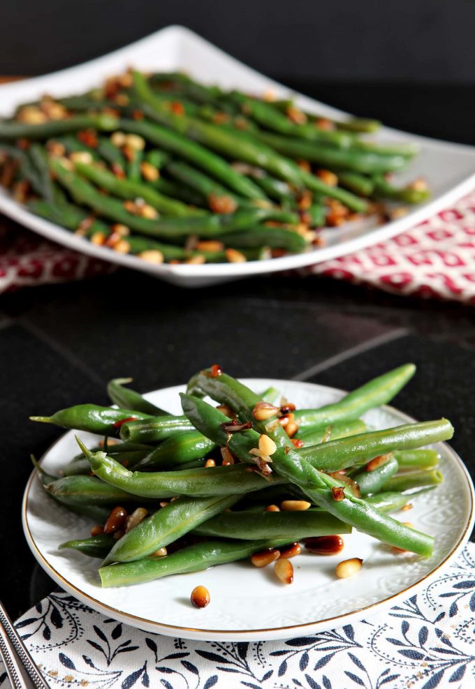 A plate of cooked green beans that are topped with roasted pine nuts. 