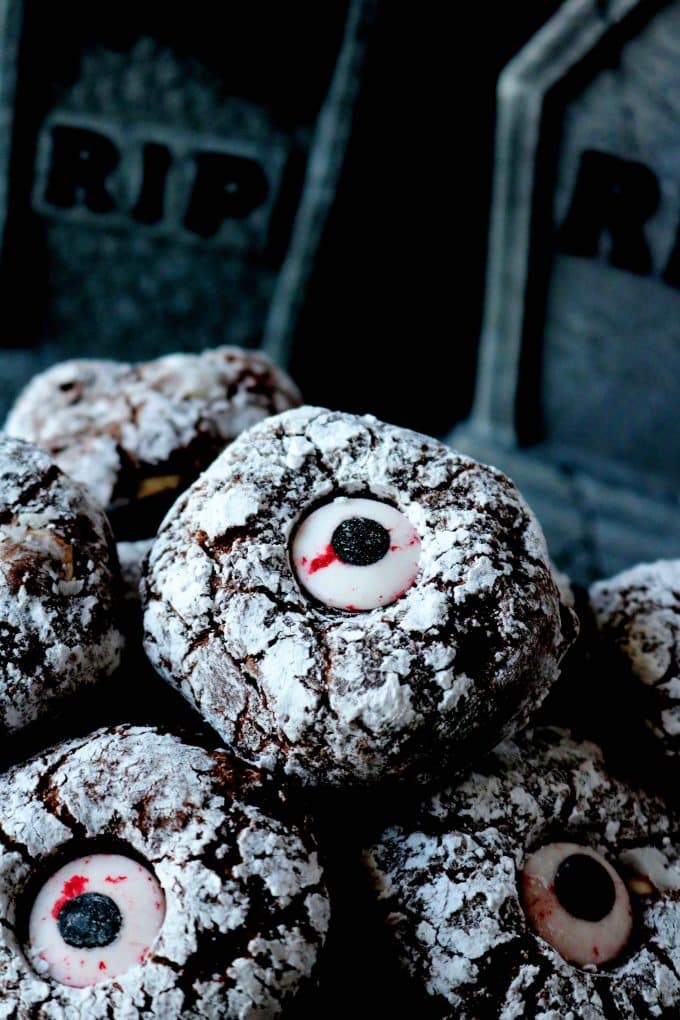 Chocolate crinkle cookies dusted with powdered sugar with a candy eye in the center. In the background are small cemetery plaques that say RIP. 