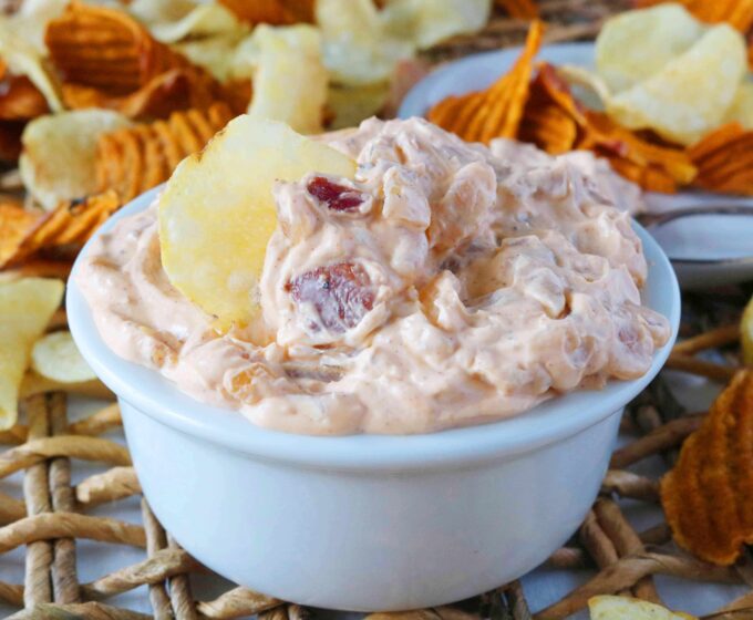 French Onion Dip with Caramelized Onion and Bacon in a small white bowl sitting on top of a woven placemat. Sweet potato and lays chips are scattered around the dip. 