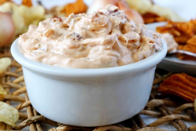 A close up shot of French Onion Dip with Caramelized Onion and Bacon in a small white bowl. In the background there are sweet potato and lays chips scattered around the bowl. 