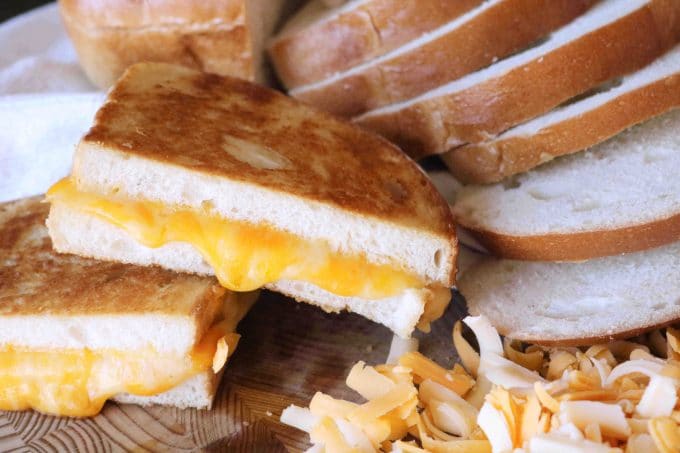 Two halves of grilled cheese sandwiches that are stacked on top of each other. Lying next to the sandwiches are slices of white bread and shredded havarti and cheddar cheeses. 
