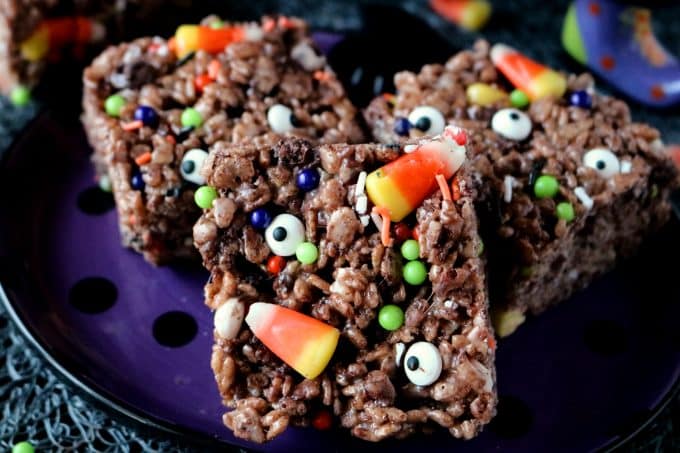 Three cut squares of Halloween Cocoa Krispie Treats topped with candy corn, eye candies, and festive sprinkles. The treats are sitting on top of a purple and black polka dot plate. 