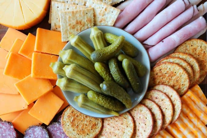 An overhead shot of pickled okra and small pickles in the center of a meat and cheese spread.