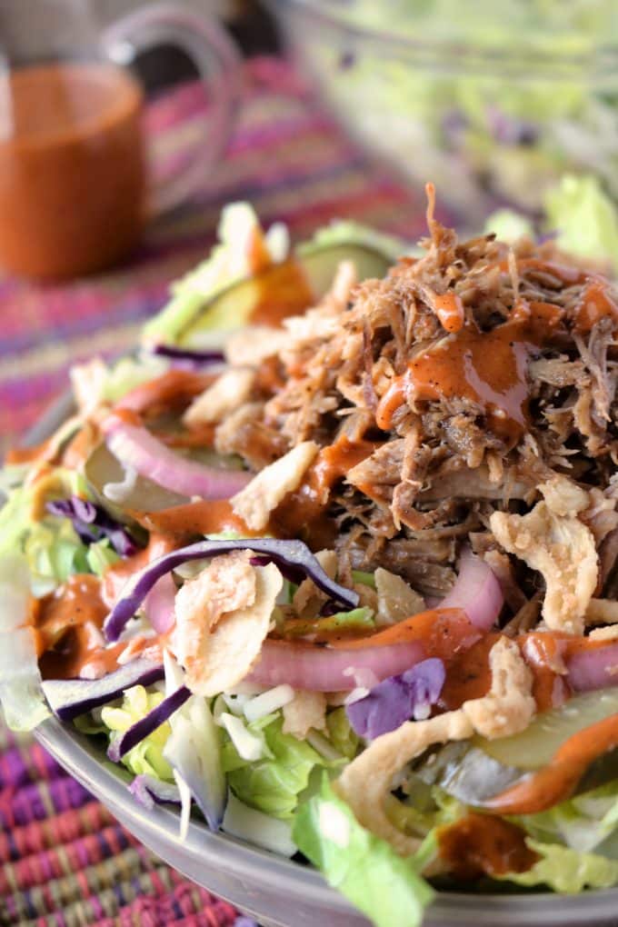 A bowl of shredded lettuce, cabbage, french fried onions, and a tangy barbecue dressing all sitting on a festive placemat. In the background is a small pitcher of salad dressing. 