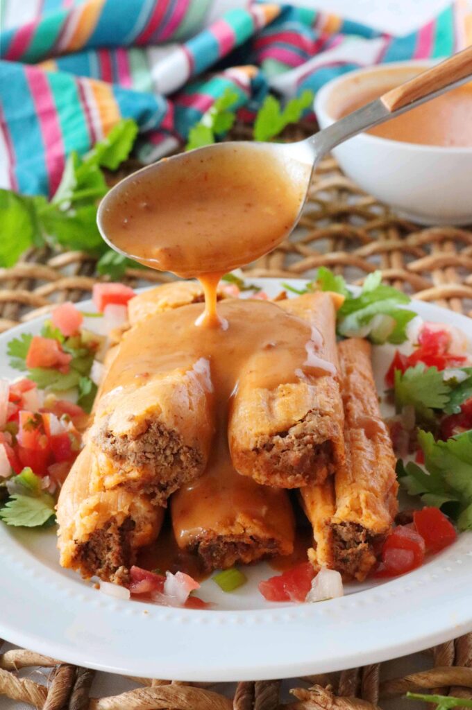 A spoonful of tamale sauce pouring over a plate of stacked tamales surrounded by pico de gallo and cilantro.