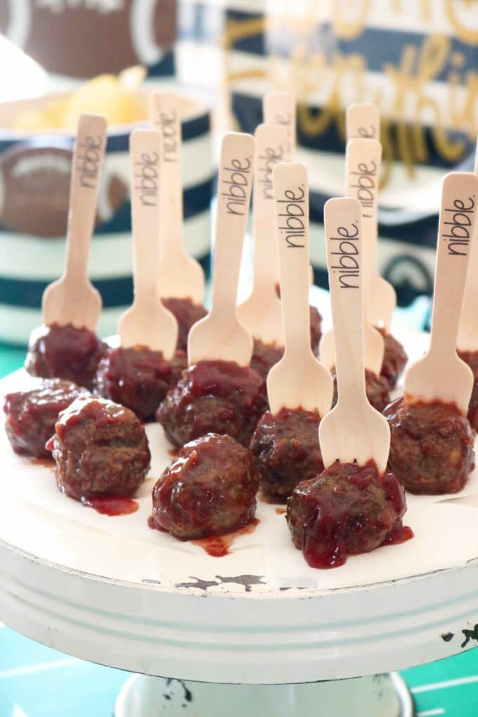 Ground beef meatballs, covered in a sweet and spicy chipotle-raspberry barbecue sauce. The meatballs are sitting on a white cake stand with wood forks that say nibble on the ends that are stuck in the meatballs. 