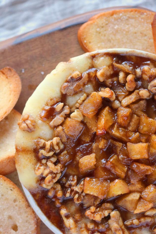 An overhead shot of baked brie topped with carmalized apples and walnuts. The small bowl of baked brie is sitting on top of a wood cutting board surrounded by toasted slices of bread. 