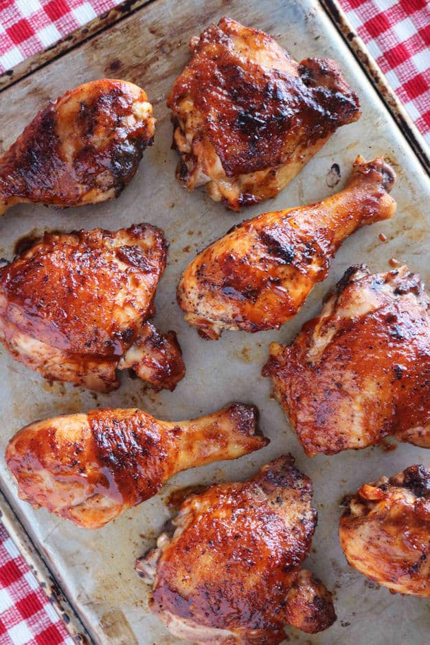 Grilled BBQ chicken legs and thighs on a baking sheet. In the left hand corner of the image is a red and white checkered tablecloth. 