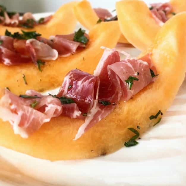 Slices of cantaloupe with thinly sliced proscuitto in the center of the cantaloupe. The cantaloupe slices are sprinkled with basil and sitting on a white serving dish. 