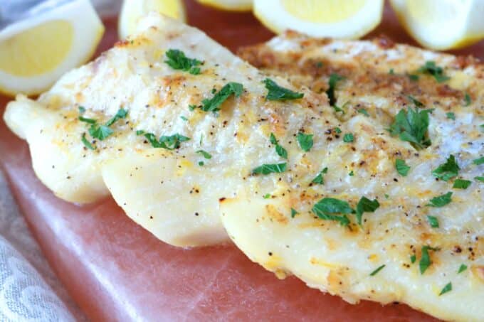  A filet of roasted cod sprinkled with fresh parsley placed on a salt block. At the top of the image are lemon quarters. 