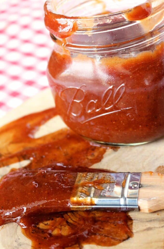 A jar of BBQ sauce sitting on top of a wooden cutting board. In front of the jar lies a basting brush covered in sauce. In the background of the image is a red and white checkered tablecloth. 