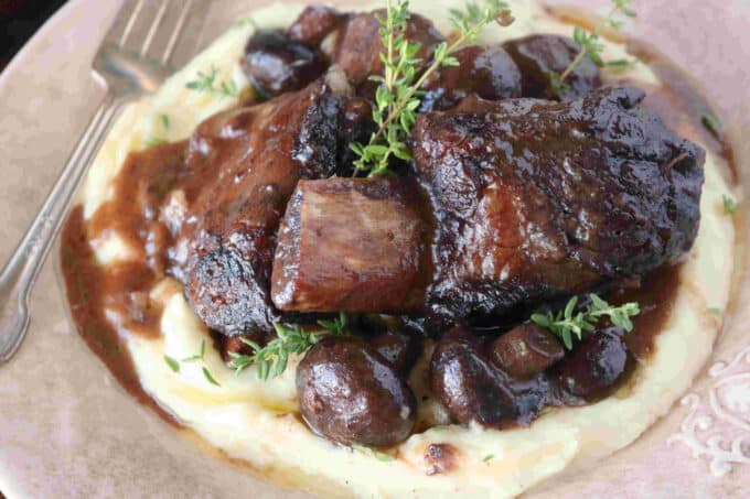 An overhead shot of Braised Short Ribs with mushrooms sitting on top of polenta. A silver fork is lying flat on the plate next to the polenta. 