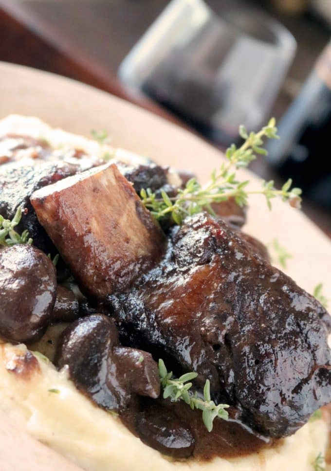 Short ribs braised in a red wine sauce sitting on top of polenta and mushrooms. Fresh thyme sprigs are scattered throughout the dish. and fresh herbs.