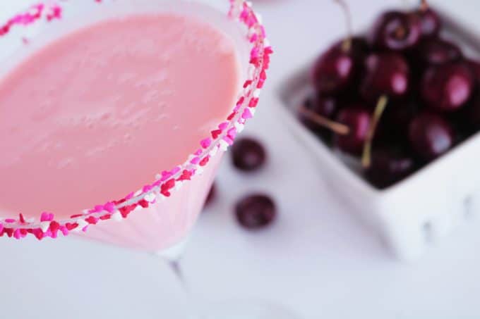 An overhead shot of a martini glass full of a cherry cheesecake cocktail rimmed with red, white and pink heart sprinkles. In the background is white basket of fresh dark cherries. 