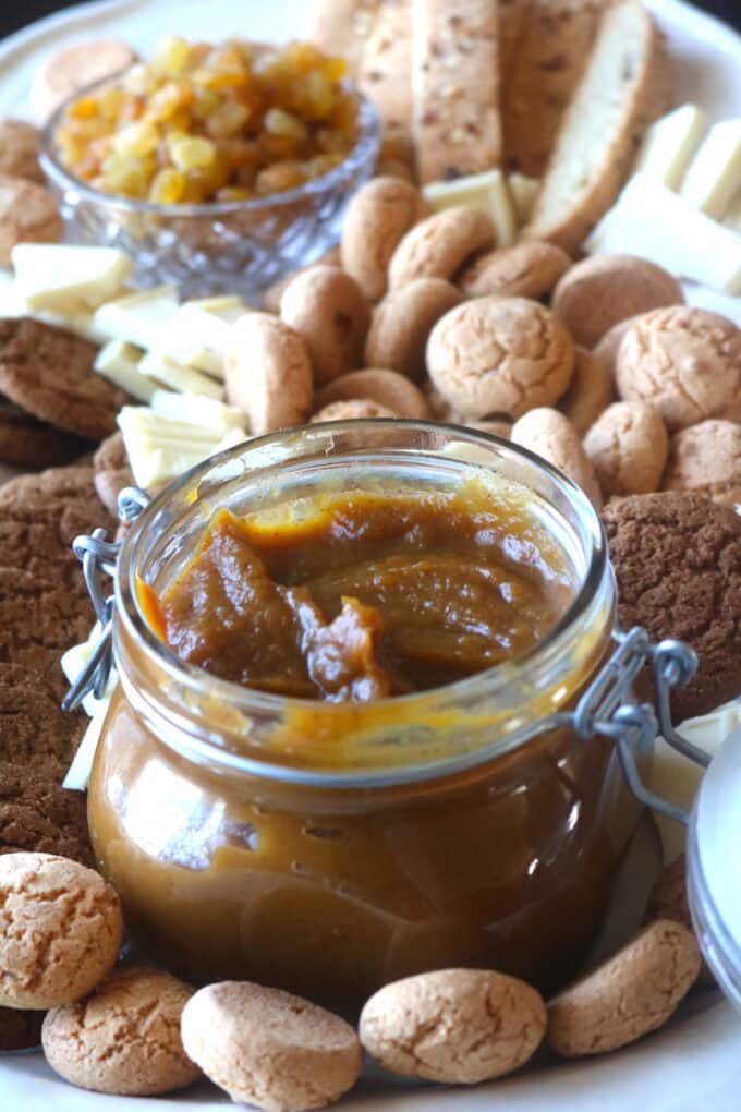 A short jar of apple pumpkin butter sitting on a white serving dish surrounded by small round cookies. In the backgroud is a small glass bowl of golden raisins along with an assortment of other cookies. 