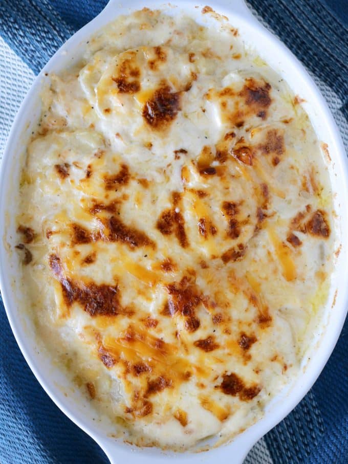 An oval serving dish full of baked creamy Scalloped Potatoes sitting on top of a blue napkin. 