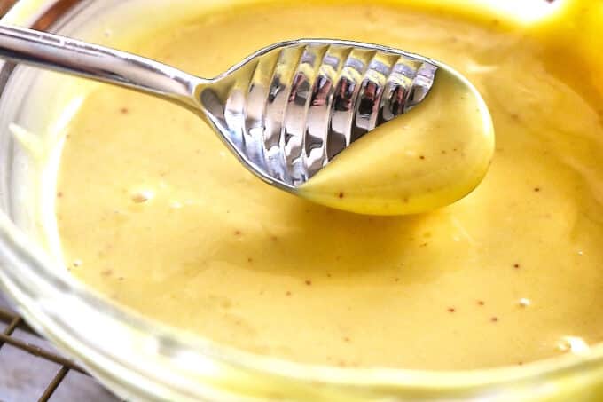 A silver honey spoon that is being held over a bowl of the honey mustard sauce. 