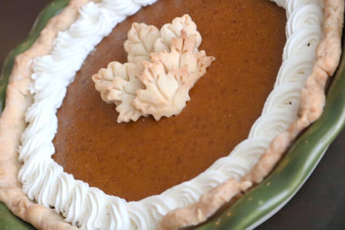 A perfect, homemade pumpkin pie that was baked in a green ceramic pie dish. Homemade whipped cream borders the outside of the pie and in the center are little leaves cut from pie crust. 