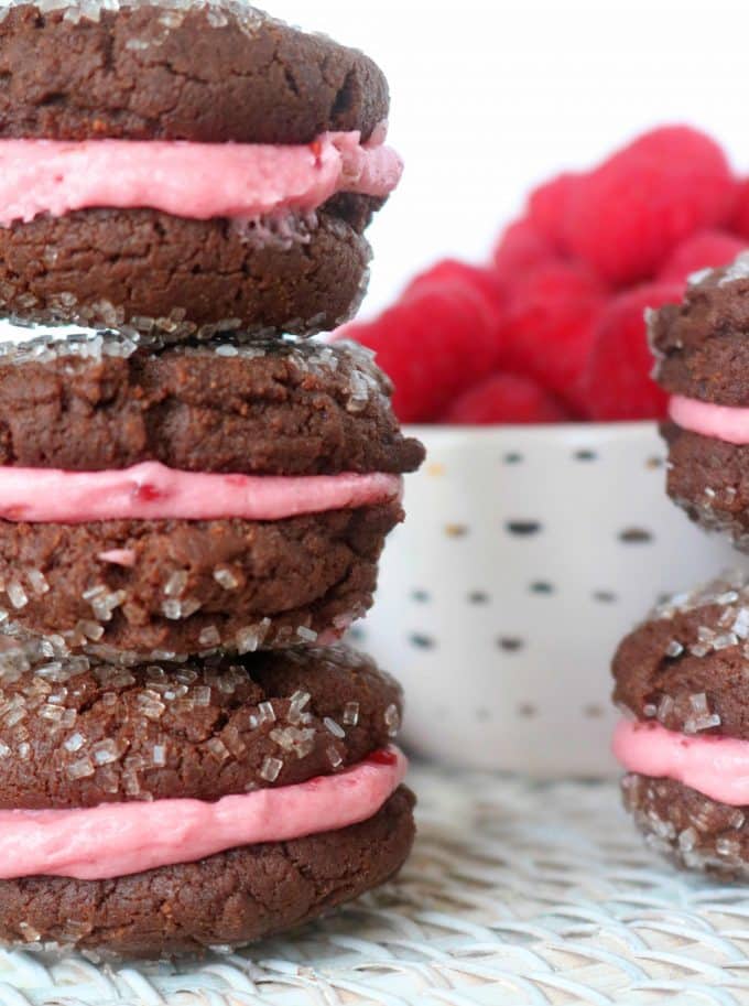 Chocolate Sandwich Cookies stacked together with a red raspberry filling. In the background are fresh raspberries in a white bowl. 