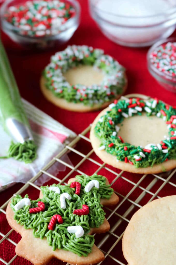 On the bottom left hand corner is a Christmas tree cookie with green icing and solid red and white candy cane sprinkles. Sitting about the tree cookie is a wreath sugar cookies with a ring of green icing and solid green, red and white sprinkles. These cookies are sitting on top of a wire cooling rack placed on a red tablecloth. Off to the side is a piping bag of green icing, a couple of glass bowls of sprinkles. 